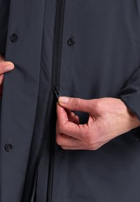 Waterdichte zwarte jas met een ritssluiting en drukknoopjes; handen zijn te zien die de rits aanpassen; gladde textuur en aansluitend ontwerp.