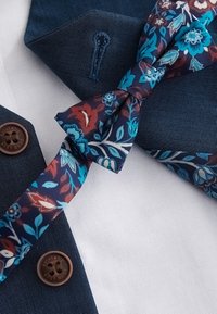 Blue textured blazer with brown buttons and matching buttonhole, paired with a blue and red floral patterned bow tie on white fabric.