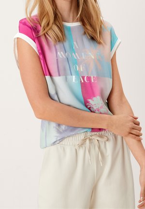 Nainen, joka pukeutunut värikkääseen hihattomaan toppiin, jossa lukee "A MOMENT OF PEACE" sekä vaalean värisiin, narulla kiristettäviin housuihin, kädet kevyesti ristissä.