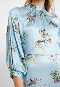 Robe en satin bleu clair ornée de motifs floraux de différentes couleurs, avec un col haut et un design à manches bouffantes. Texture lisse dans l'ensemble.