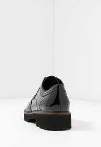 Sorte patenterede lædersko med brogue-detaljer, rund tå, tyk sort sål og kontrasterende brun syning langs kanten.