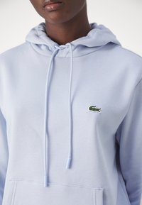 Sweatshirt de algodão azul claro com um bolso tipo canguru, capuz com cordão e um logotipo de crocodilo bordado em verde no peito. Textura suave.