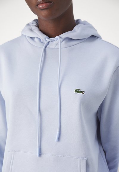 Haut en coton bleu clair avec une poche kangourou, un capuchon à cordon de serrage, et un logo de crocodile vert brodé sur la poitrine. Texture lisse.