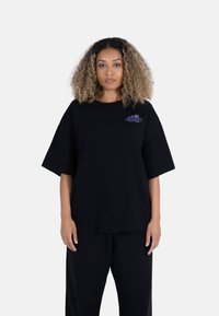 Svart oversized t-skjorte laget av myk bomull, med korte ermer og en lilla logo på venstre bryst. Kombinert med løse svarte bukser.