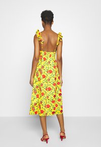 Vestido amarillo con estampado floral, tirantes ondulados y cintura elástica. La espalda está descubierta y tiene un diseño de longitud media. Combinado con sandalias rojas.