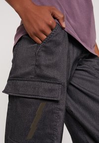 Harmaat työhaalarit, joissa suuri sivutasku, sileä tekstuuri ja taskussa huomaamaton yksityiskohta. Yhdistetty violettiin paitaan.