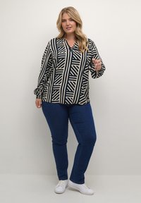 Langærmet, knaplukket bluse med et geometrisk sort og hvidt mønster, parret med blå jeans og hvide sneakers.