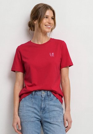 Młoda kobieta uśmiecha się, nosi czerwoną koszulkę z napisem „LOVE” na piersi i niebieskie, wysokowciskowe dżinsy, stoi na tle jednolitej jasnej ściany.