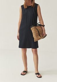 Femme portant une robe noire sans manches et des sandales noires, tenant un sac à main tissé beige et noir contre un fond neutre.