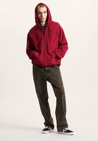 Röd zip-up hoodie med dragsko, framfickor, bärs över en mörkgrön skjorta, kombinerad med olivgröna byxor och svarta sneakers.