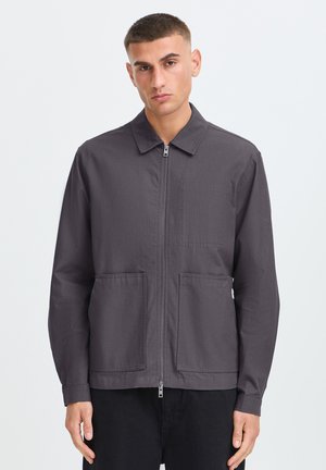 Junger Mann trägt eine dunkelgraue Jacke mit Reißverschluss, Kragen und zwei vorderen Taschen, steht vor einem schlichten hellen Hintergrund.