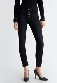 Jeans neri skinny con vita alta, dotati di chiusura frontale a cinque bottoni e tasche discrete, abbinati a scarpe con tacco alto scintillanti.