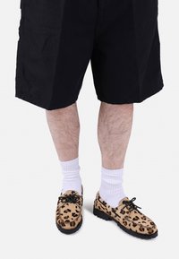 Leopardenmuster, Wildlederschuhe mit schwarzen Sohlen, mit abgerundeten Zehen und Schnürsenkeln, getragen mit weißen, gerippten Knöchelsocken und schwarzen Shorts.