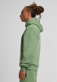 Sweat à capuche vert avec une coupe décontractée, doté d'une grande capuche et de poignets côtelés. Fabriqué en matériau doux, il possède un discret logo sur le côté.