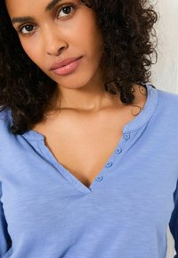 Light blue long-sleeve top with a V-neckline, featuring four small blue buttons on the placket and a soft, textured fabric.