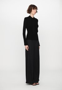 Black fitted ribbed top with a tie collar, paired with wide-leg black trousers, both in smooth fabric. Model stands in profile against white background.