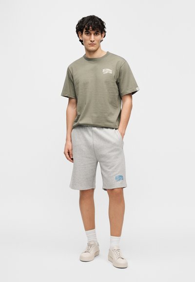 Πράσινο βαμβακερό μπλουζάκι με λογότυπο, σε συνδυασμό με γκρι βαμβακερά σορτς που έχουν μπλε γραφική λεπτομέρεια, φορεμένα με ανοιχτά αθλητικά παπούτσια και λευκές κάλτσες.