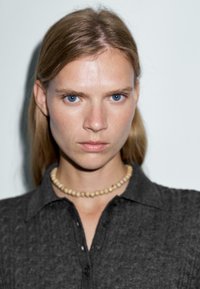 Graues Strickpolohemd mit Kragen, kombiniert mit einer kurzen, perlbesetzten Halskette. Das Model hat glattes, blondes Haar und blaue Augen. Einfache Hintergrund.
