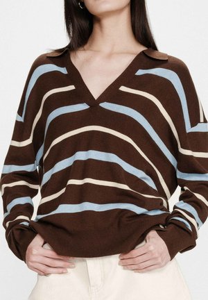 Femme portant un pull marron à col en V avec des rayures ondulées bleues et beige, associée à un pantalon écru, les mains glissées à la taille.