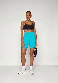 Schwarzer Sport-BH mit weißem Nike-Logo, kombiniert mit türkisfarbenen, hochgeschnittenen Radshorts und weißen Sneakers. Hält eine schwarze Wasserflasche.