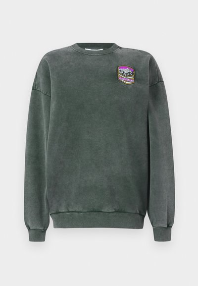 Sweatshirt verde escuro com gola redonda e punhos canelados. Apresenta um emblema com um design de montanha em roxo, branco e amarelo. Textura suave.