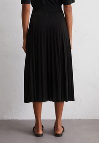 A person stands with their back to the camera, wearing a black pleated skirt and black sandals, against a neutral textured background.