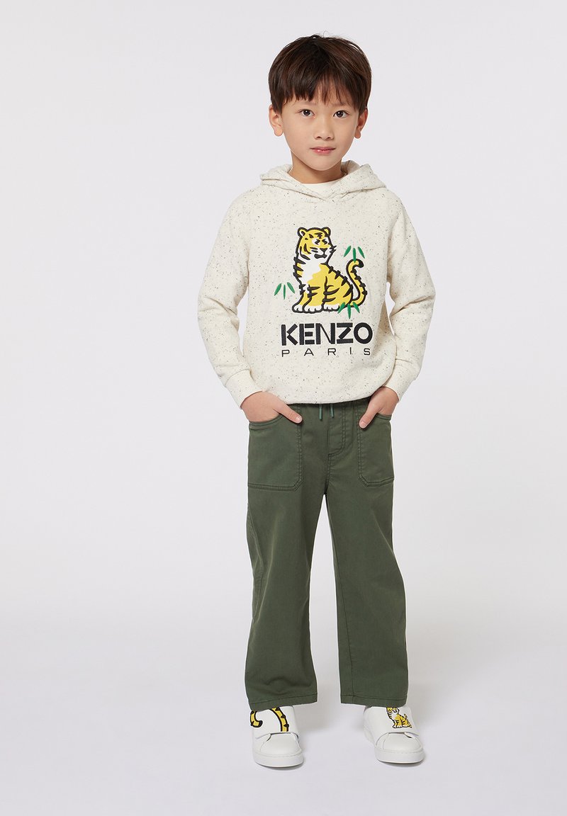 Haut à capuche crème avec un graphisme de tigre et le texte "KENZO PARIS", associé à un pantalon vert et des chaussures blanches avec un design jaune.