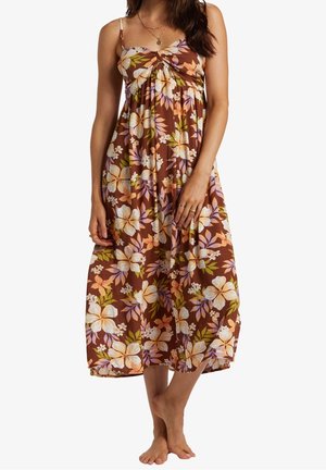 Kobieta w brązowej, bezrękawnikowej sukience midi w biało-różowo-zielony wzór kwiatowy, stojąca boso na jednolitym tle.