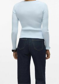 Personne portant un haut à manches longues côtelé bleu clair et un jean en denim bleu foncé, debout de dos sur un fond blanc.