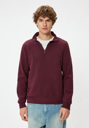 Sudadera de algodón burdeos con cuello de media cremallera, puños y dobladillo acanalados, y un corte relajado, combinada con jeans de mezclilla azul claro.