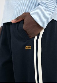 Marineblaue Jogginghose mit weißen Seitestreifen, ausgestattet mit einem gestickten gelben Wellenlogo. Die Hand des Models ist in der Tasche, kombiniert mit einem hellblauen gestreiften Hemd.