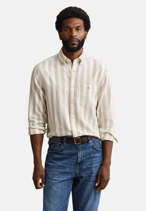 Mann mit Bart trägt beige-weiß gestreiftes Langarmhemd, blaue Jeans und einen braunen Gürtel vor einem einfarbigen Hintergrund.