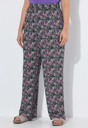 Vrouw draagt losse, hooggetailleerde broek met een geometrisch patroon in zwart, wit, roze en groen, gecombineerd met beige sandalen en een paarse top.