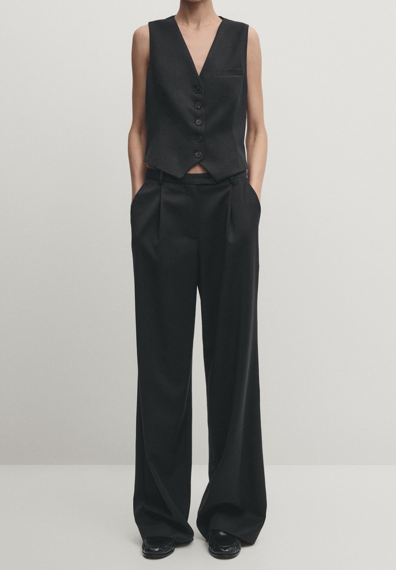 Ärmlös svart knäppt väst i kombination med vida svarta byxor och svarta skor på en stående person mot en enfärgad bakgrund.