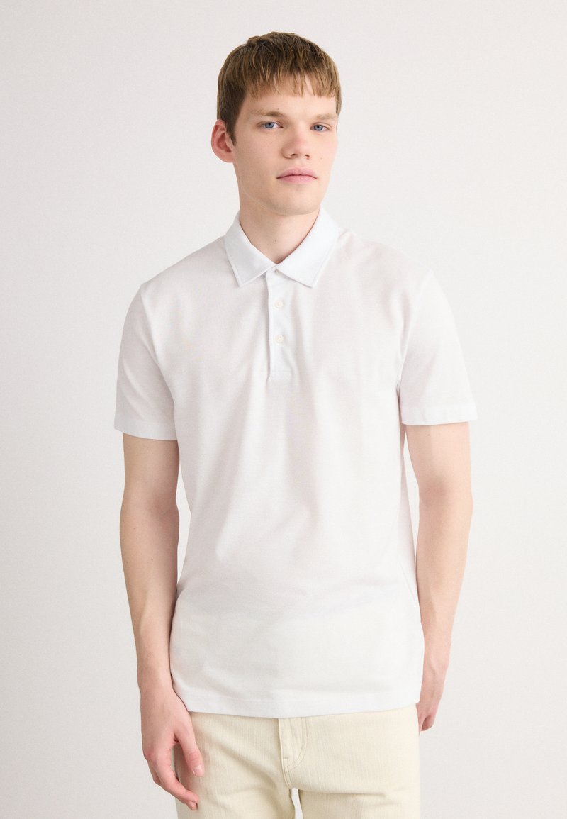 Mlad moški s svetlimi lasmi, oblečen v belo majico s kratkimi rokavi in bež hlačami, stoji ob lahkem enotnem ozadju.