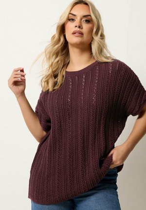Blonde Frau trägt einen locker sitzenden, kurzärmligen, dunkelvioletten Strickpullover und blaue Jeans, posiert mit einer Hand nahe am Gesicht und der anderen in der Hosentasche.