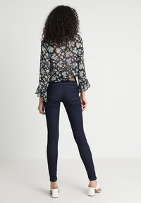 Blusa negra transparente con estampado floral y mangas con volantes, combinada con jeans ajustados de color azul oscuro y zapatos blancos de tacón cuadrado. Vista trasera.