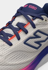 Κοντινό πλάνο λευκού πλεκτού παπουτσιού τρεξίματος New Balance 1080 με ναυτικά κορδόνια, ναυτικό λογότυπο, ροζ λεπτομέρειες και σόλα με βαθμιαία απόχρωση από μωβ σε ροζ.