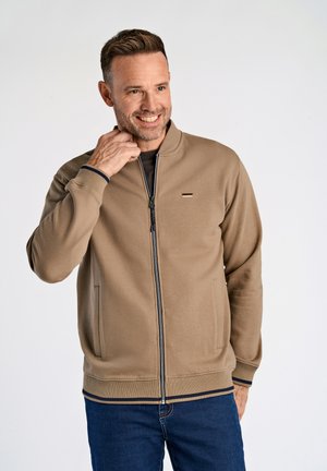 Chaqueta beige con cremallera, cuello alto, puños y dobladillo acanalados. Presenta un pequeño logo en el pecho y dos bolsillos laterales.