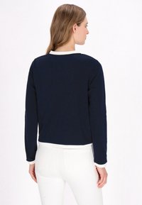 Navy long-sleeve top with white collar and cuffs, made from a smooth fabric. Model wears white jeans, showing a clean, fitted design.