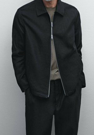 Homme portant une veste à fermeture éclair noire par-dessus une chemise vert olive, les mains dans les poches, sur un fond clair uni.