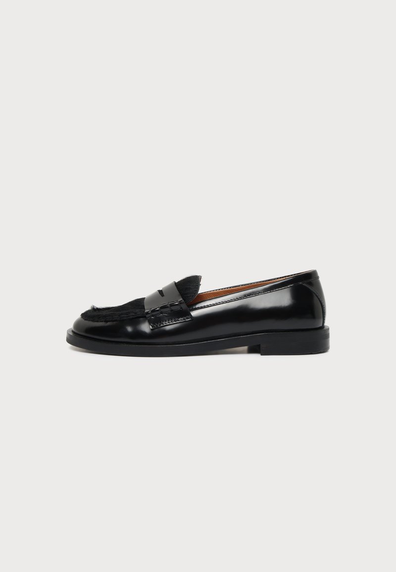 Mocassin en cuir noir avec un petit talon et un détail en fourrure noire sur le dessus, montré de côté extérieur sur fond blanc.