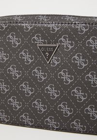 Bolso negro texturizado con un patrón de diamantes y el logo de Guess en plata en un acento metálico triangular. Cierre de cremallera, diseño compacto.