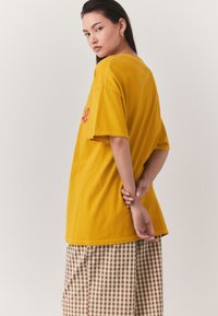 Maglietta oversize di colore giallo senape, con maniche corte e orlo arrotondato. Abbinata a pantaloni larghi a quadri marroni e crema.