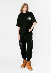 Camisa negra de gran tamaño con estampado gráfico, combinada con pantalones joggers negros con bolsillos con cremallera y botas marrones con cordones. Look sencillo y casual.