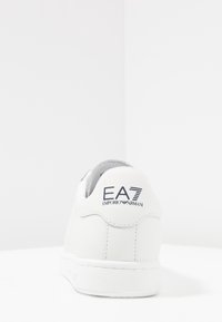White leather sneaker with low-top design. Features branded heel tab with "EA7 Emporio Armani." Smooth texture and round toe shape.