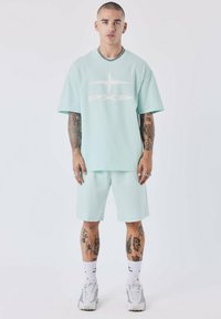 Ensemble t-shirt à manches courtes vert menthe clair et short avec logo "PXP" blanc. Le modèle porte des chaussettes blanches et des baskets grises, affichant des tatouages sur les bras.