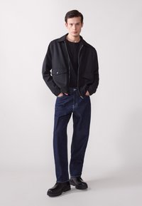 Chaqueta negra de cremallera con dos bolsillos frontales sobre una camiseta negra, combinada con jeans de mezclilla oscuros y suéteres de plataforma negras.