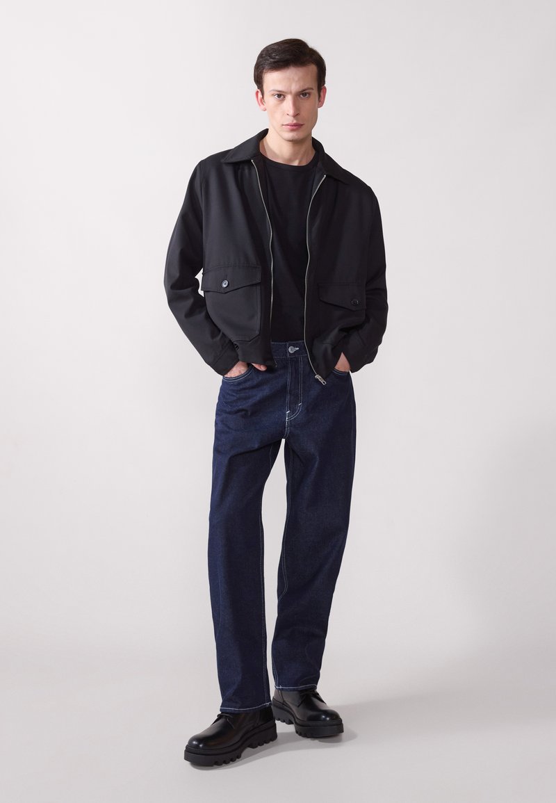 Chaqueta negra de cremallera con dos bolsillos frontales sobre una camiseta negra, combinada con jeans de mezclilla oscuros y suéteres de plataforma negras.