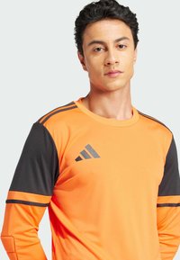 Helles orange langärmliges Sportshirt mit schwarzen Akzenten und grauem Dreistreifen-Logo auf der linken Brust und den Ärmeln, aus leichtem Stoff hergestellt.